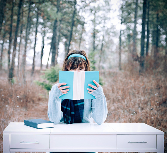 Creative photo of woman with book in front of her. She is in a field sitting at a white desk.
