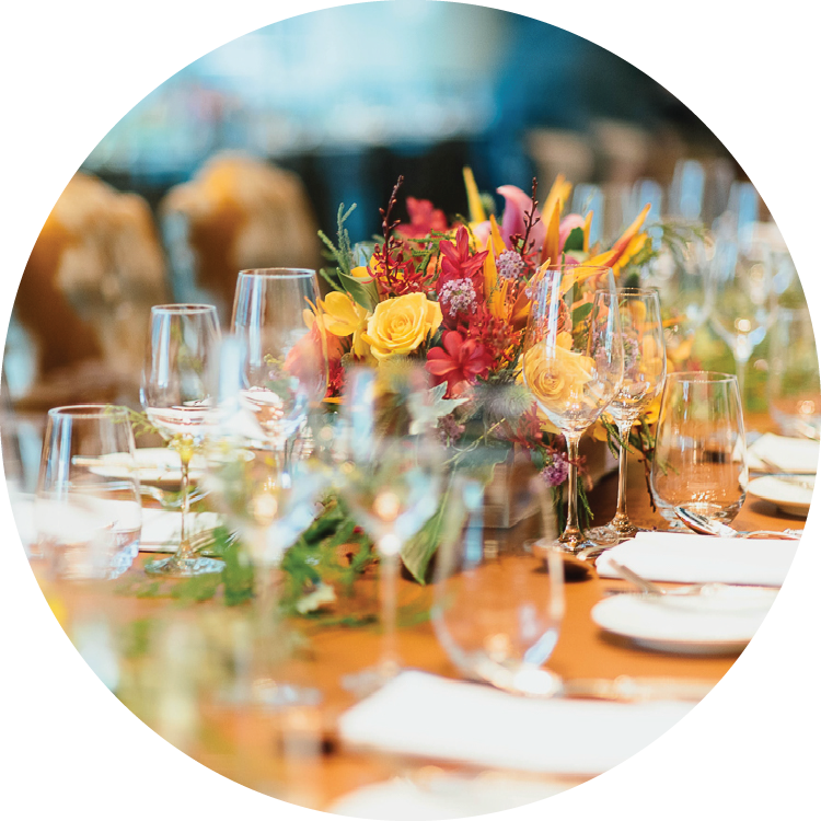 Formal place settings are seen down a long table including three glasses, flatware, a small plate, and napkin at each place. Greenery is strewn down the center of the table with a yellow and red floral arrangement in the center.