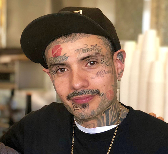 Man with tattoos, black shirt, and black baseball cap poses for a photo 