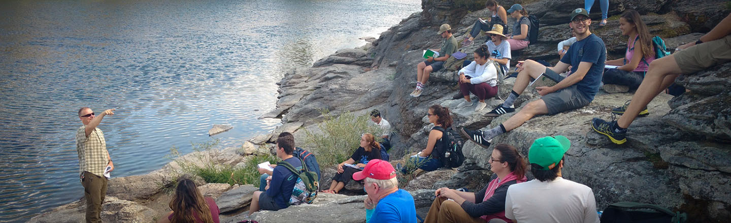 Students sitting on a rock hillside with instructor near waters edge.