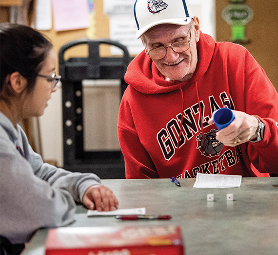 An elderly man wearing a Gonzaga hoodie plays a dice game with a student.  