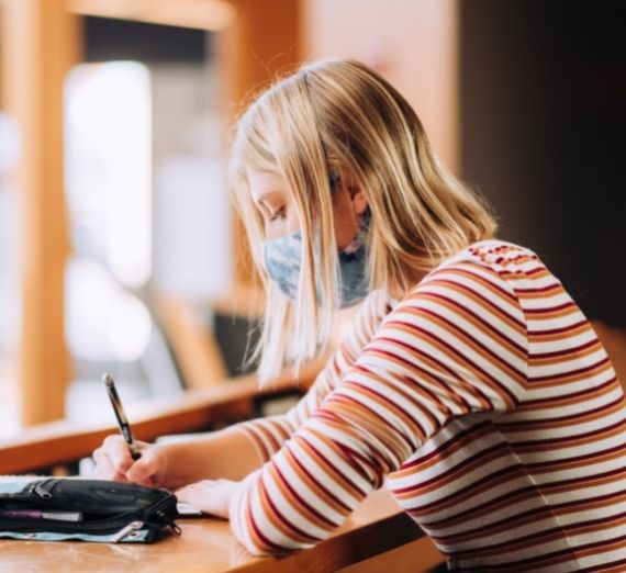 A student sitting indoors writing. She is wearing a blue mask and a red and white striped shirt. Her hair is blonde and the background is blurred. 
