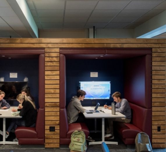 This is a photo of students working in the Rosauer Center's Active Learning Center. To the right, two students are working in a booth facing each other, with backpacks at their feet. The flat screen TV behind them is on. To the left, there is another booth not fully seen with at least two students working. The TV is off behind them. 