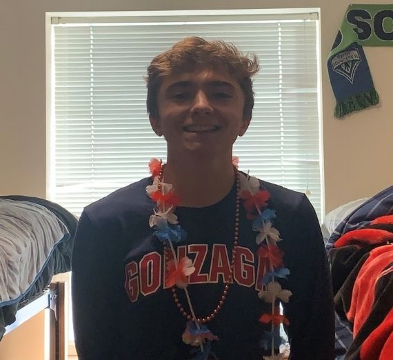 A current student smiling in his residence hall room. He is wearing a navy long sleeved shirt with GONZAGA written in red letters. He is wearing a lei. 