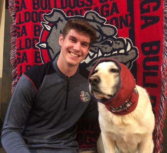 A current student sitting with a dog. The student is wearing a Gonzaga pullover and the dog is wearing a Gonzaga neckwarmer/hat. The background is a Gonzaga Bulldogs blanket. 