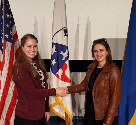 Two women smiling and shaking hands in front of three flags 