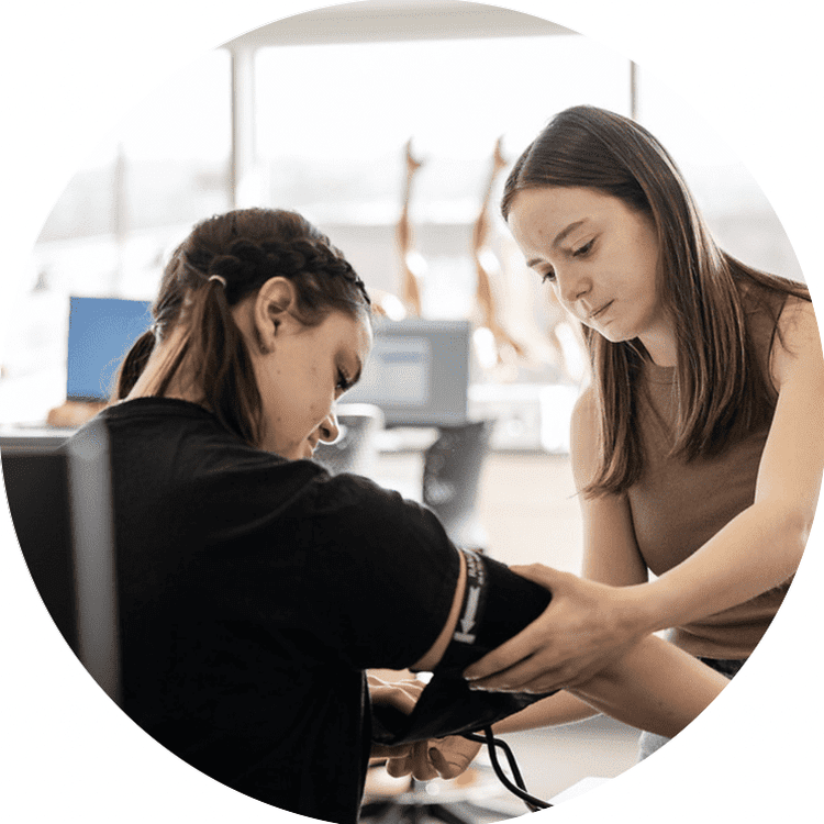 A young woman assists another woman by measuring her blood pressure in a clinical or educational environment. The setting appears bright and modern, with medical or anatomical models visible in the background. A young woman assists another woman by measuring her blood pressure in a clinical or educational environment. The setting appears bright and modern, with medical or anatomical models visible in the background.