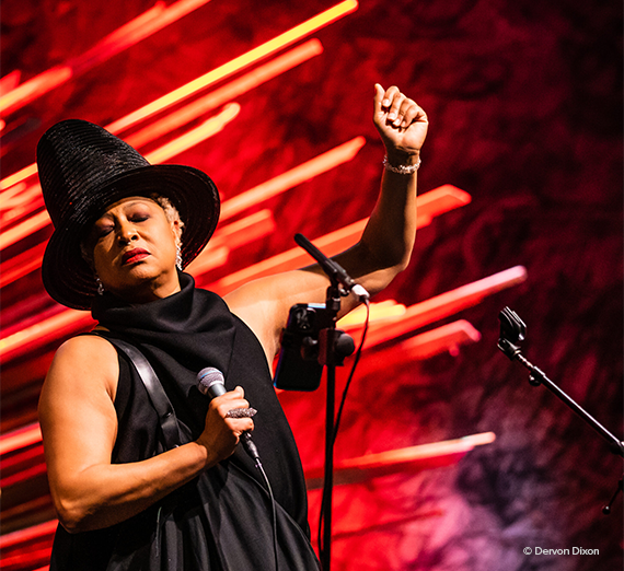 Ms. Lisa Fischer on stage against a red and orange background wearing a black hat and black dress holding a microphone. 
