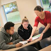 Teacher pointing at a book with two students sittin in chairs looking at the book.