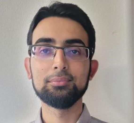 man with dark hair, facial hair, and glasses standing in front of white background