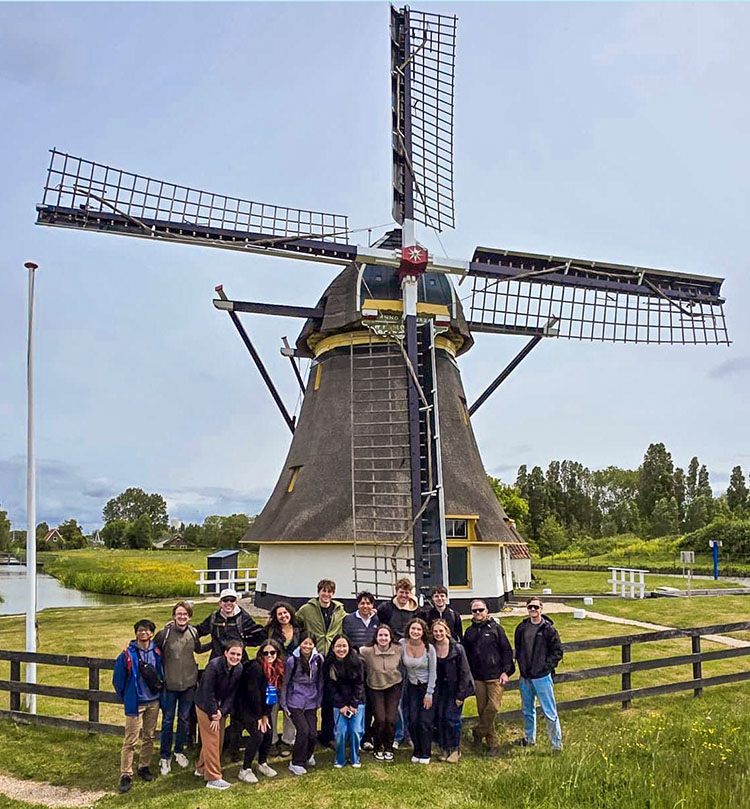 18 students and other adults stand at the base of a large, traditional Dutch windmill