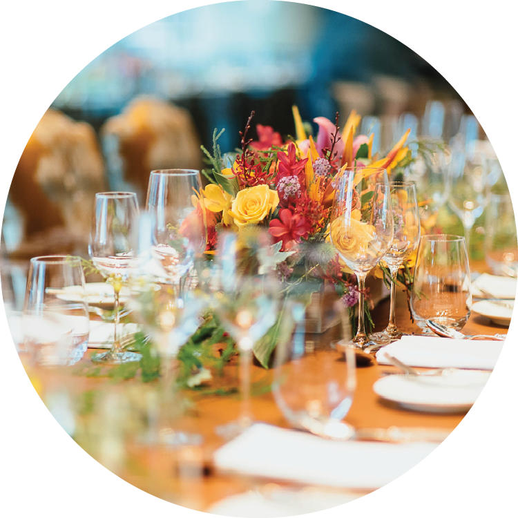 Formal place settings are seen down a long table including three glasses, flatware, a small plate, and napkin at each place. Greenery is strewn down the center of the table with a yellow and red floral arrangement in the center. Formal place settings are seen down a long table including three glasses, flatware, a small plate, and napkin at each place. Greenery is strewn down the center of the table with a yellow and red floral arrangement in the center.