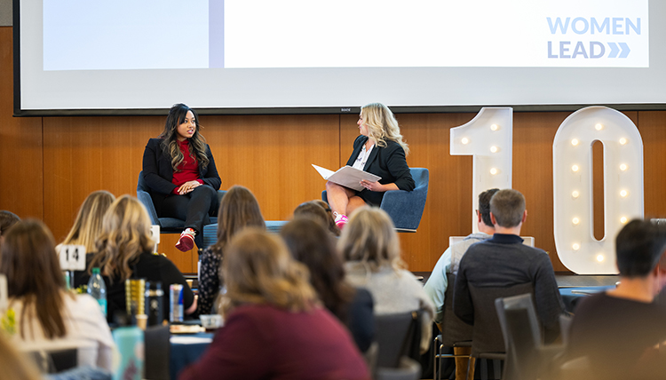 Two women converse on a stage while a seated audience listens Two women converse on a stage while a seated audience listens