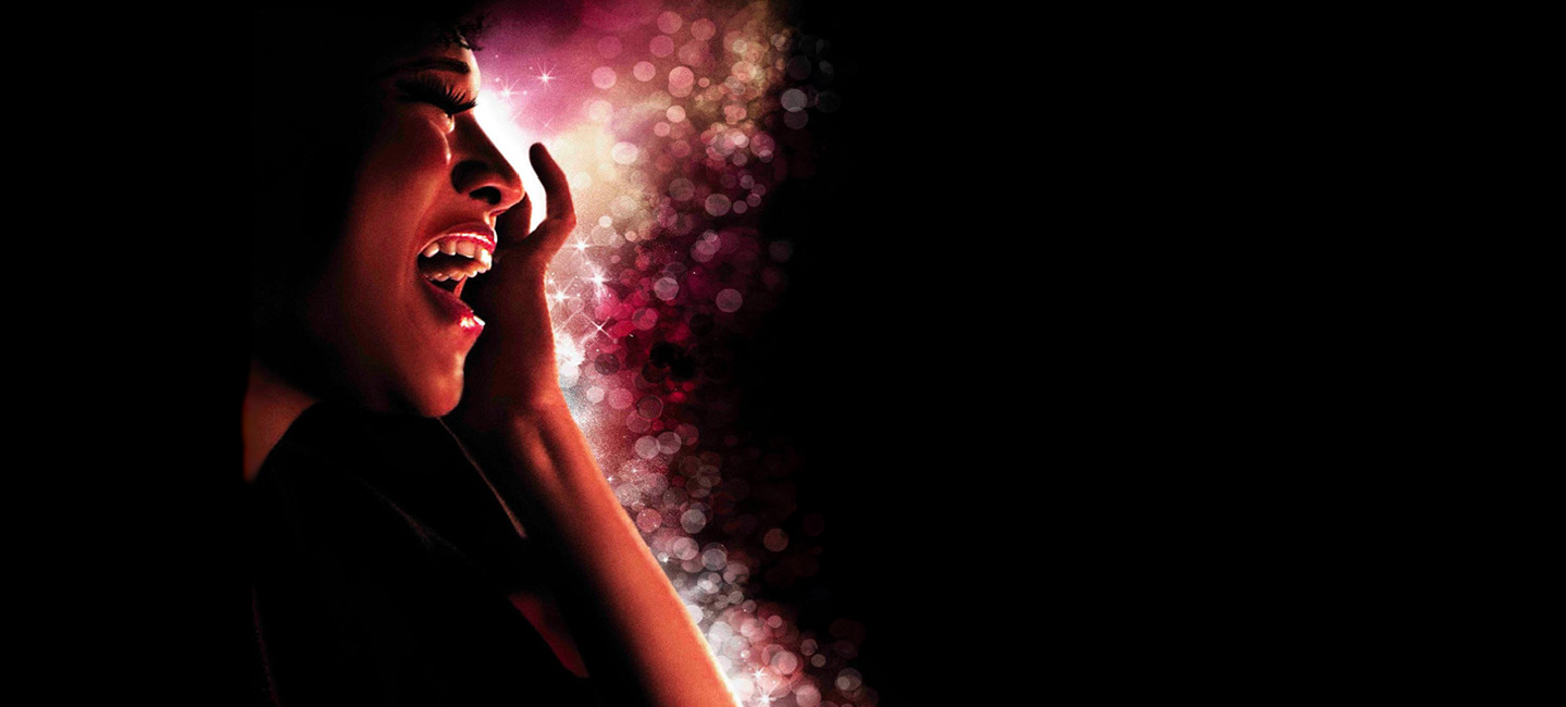 A close up of an artist's face as they sing passionately, against a sparkling pink and black background.