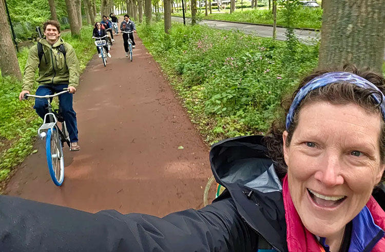 A woman's face smiles in the lower right hand corner. Over her shoulder is a view of students riding bicycles on a forested trail.