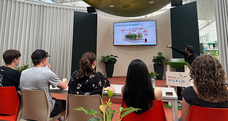 Five students sit in a conference room looking at a man pointing to the screen that says Technology in the field lab over an illustration of plants in a greenhouse