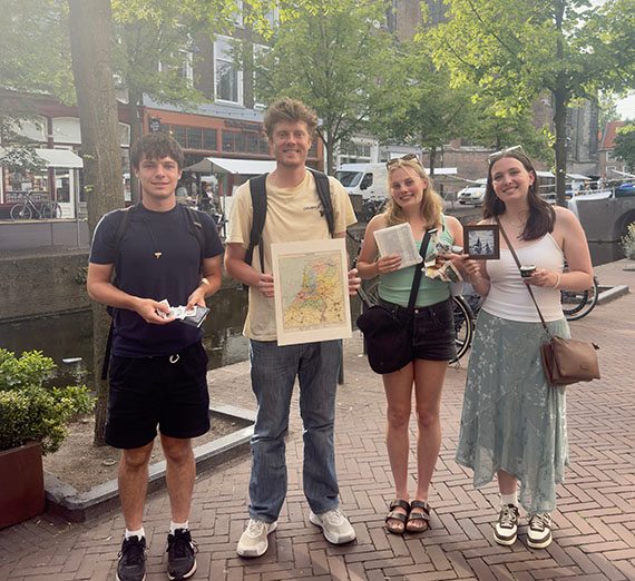 Four young adults stand in a row on a paved walkway, each holding a paper, book, or picture in frame