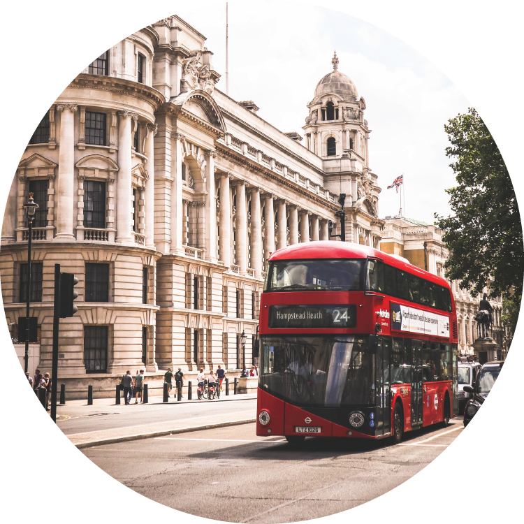 A red double-decker bus drives past the front of a long, white columned building in downtown London. A red double-decker bus drives past the front of a long, white columned building in downtown London.