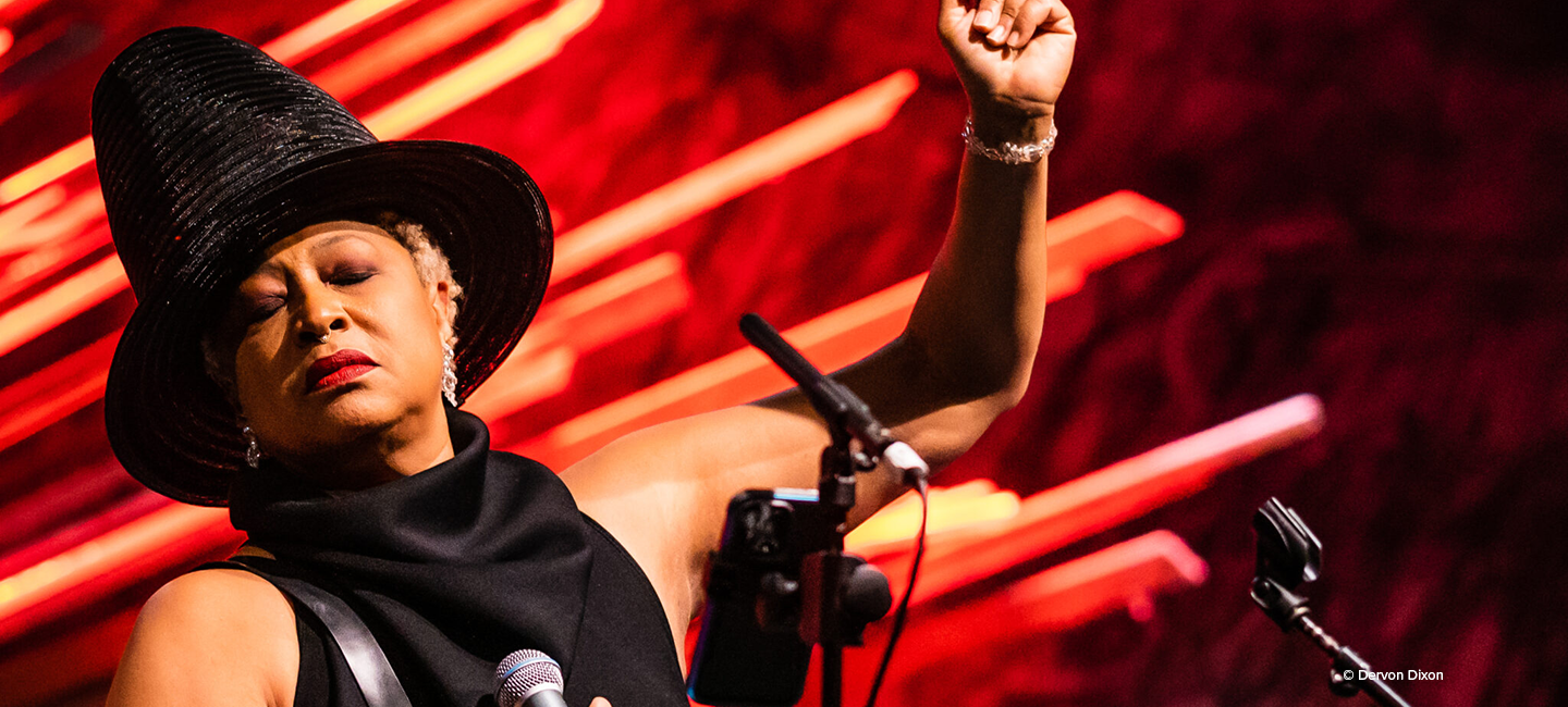 Ms. Lisa Fischer on stage against a red and orange background wearing a black hat and black dress holding a microphone. 