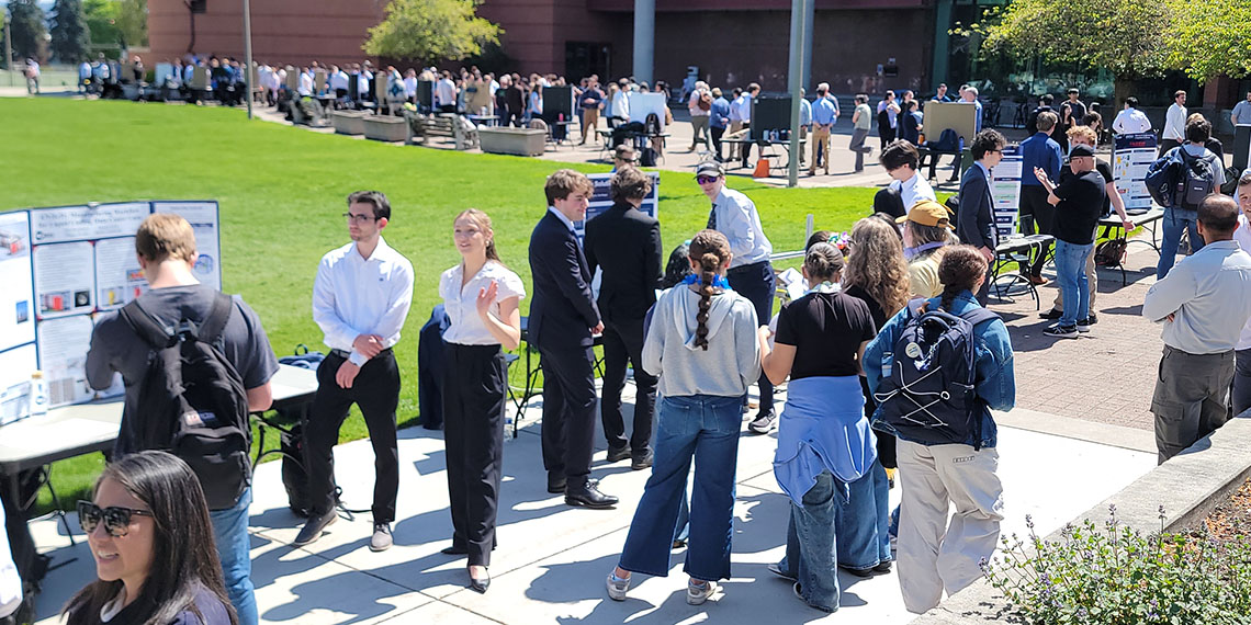 A hundred people gather at tables spaced out around the edge of a square lawn on a sunny day.