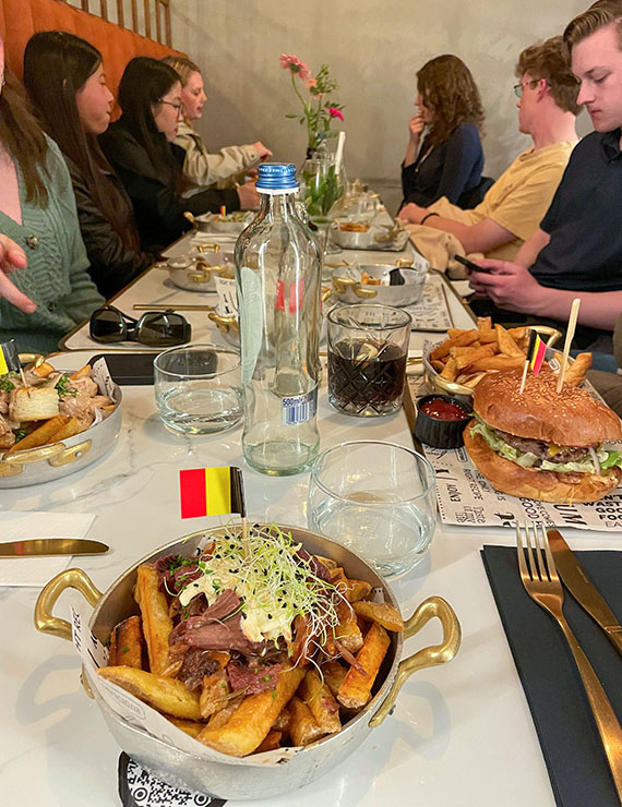 several young adults sit around a long table with large plates of food, most with a small paper Belgian flag of black, yellow and red vertical bands