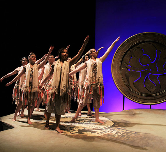 Performers on stage in fringed costumes, extending arms in a choreographed dance under purple lighting. Performers on stage in fringed costumes, extending arms in a choreographed dance under purple lighting.
