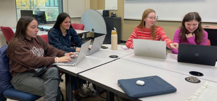 Four students working together on laptops around a conference-style table in a modern classroom with whiteboards, a projector screen, and large windows. A tablet and water bottle are on the table, highlighting a collaborative learning environment with digital tools. Four students working together on laptops around a conference-style table in a modern classroom with whiteboards, a projector screen, and large windows. A tablet and water bottle are on the table, highlighting a collaborative learning environment with digital tools.