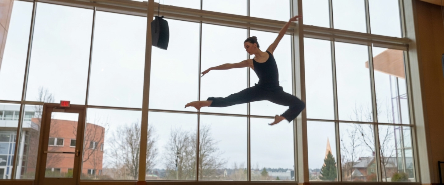 A dancer in a black leotard leaping mid-air in front of tall windows. 