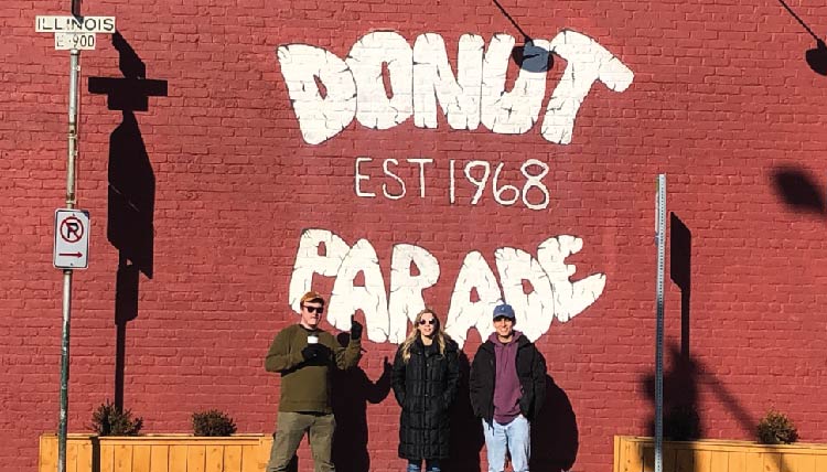 three students standing in front of a building with the words 'Donut Parade, est 1968' inscribed on it three students standing in front of a building with the words 'Donut Parade, est 1968' inscribed on it