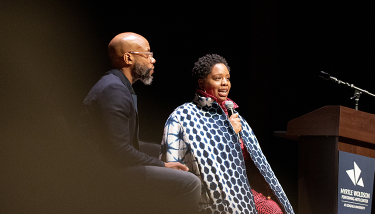 Woman with microphone talking onstage, next to man who is listening as they lead a discussion together Woman with microphone talking onstage, next to man who is listening as they lead a discussion together