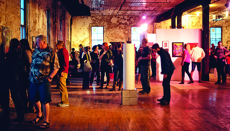 A group of spectators look at a variety of art exhibits in a brick room with wood floors. A group of spectators look at a variety of art exhibits in a brick room with wood floors.