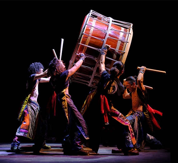 Yamato: The Drummers of Japan performers in a high-energy pose, lifting and striking a large traditional Taiko drum with wooden bachi sticks on a dark stage.