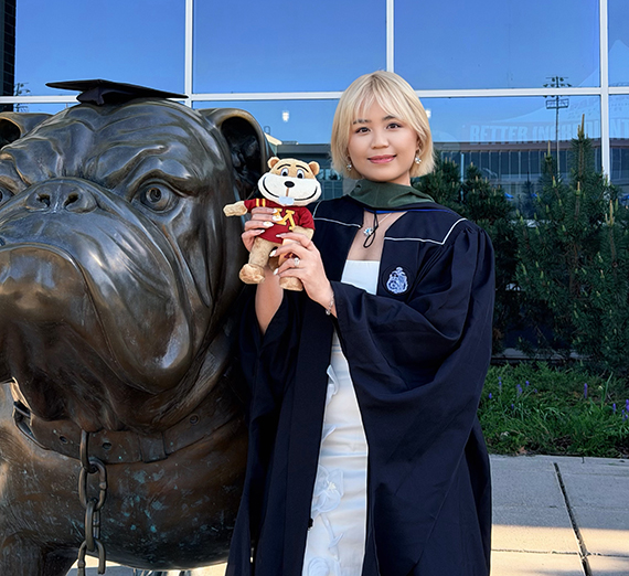 Blonde female wearing graduation robe standing next to bulldog statue and holding stuffed animal