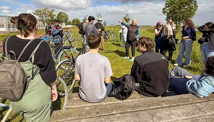 Young adults sit outdoors in grass with attention on a teacher.