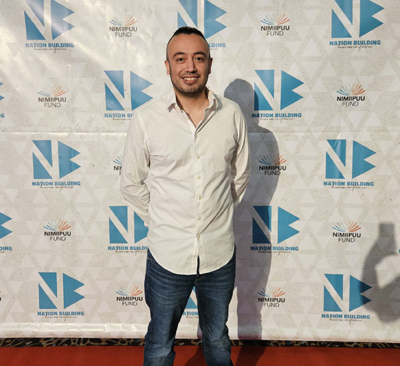 Male standing on red carpet with white and blue background. Male is wearing a white button up and jeans with short brown hair.