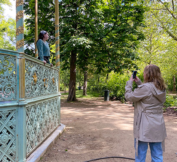 Young woman stands in an ornate bandstand while another on a forested trail takes her picture