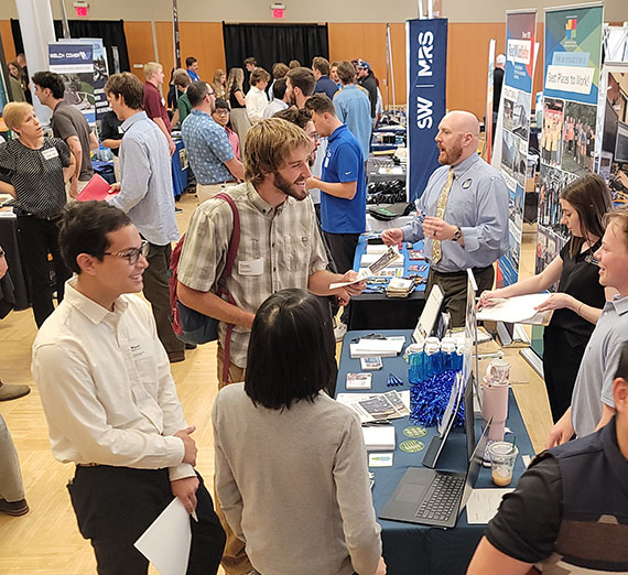A busy row of tables in an assembly hall where students and professionals talk excitedly