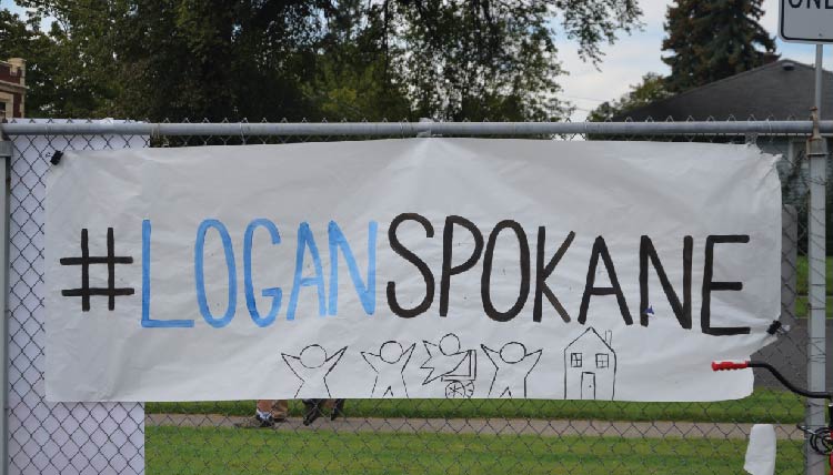 a hand-drawn sign fastened to a chain-link fence. The sign reads #LoganSpokane with a illustration of children and a house on the bottom of the sign. a hand-drawn sign fastened to a chain-link fence. The sign reads #LoganSpokane with a illustration of children and a house on the bottom of the sign.