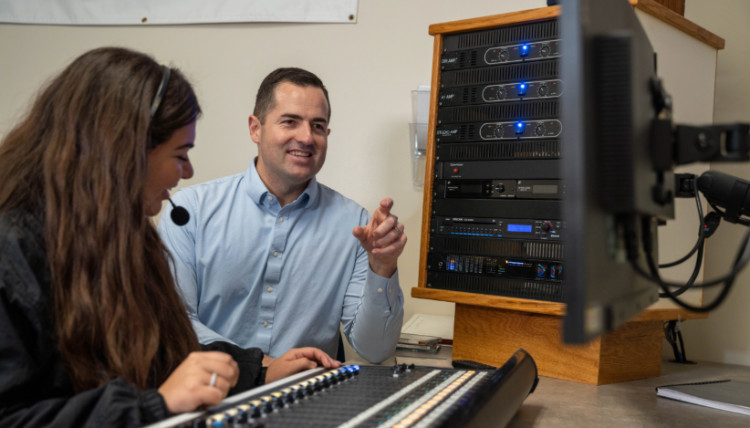 Broadcast journalism student operating an audio mixing console while a faculty member provides guidance in a university media control room equipped with professional sound and production equipment. Broadcast journalism student operating an audio mixing console while a faculty member provides guidance in a university media control room equipped with professional sound and production equipment.