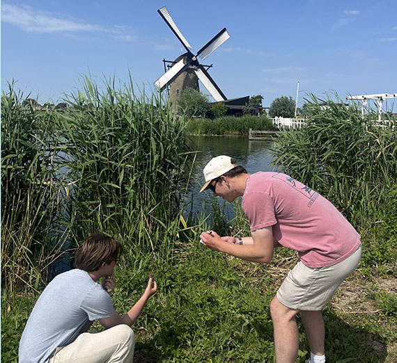 Two young men at the edge of a canal hold up pieces of dirt while the other writes down data