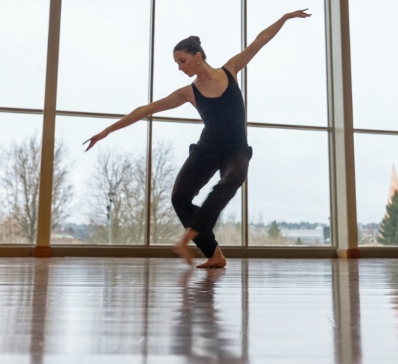 A dancer wearing a black leotard in motion in front of a large window.