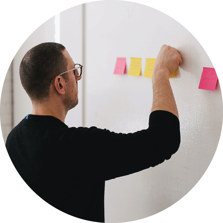 A man in a black sweater stands in front of a whiteboard and reaches up to place a colored sticky note alongside a line of yellow and pink notes. A man in a black sweater stands in front of a whiteboard and reaches up to place a colored sticky note alongside a line of yellow and pink notes.