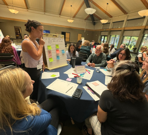 A facilitator stands at a round table listening to a smoke symposium participant share feedback A facilitator stands at a round table listening to a smoke symposium participant share feedback