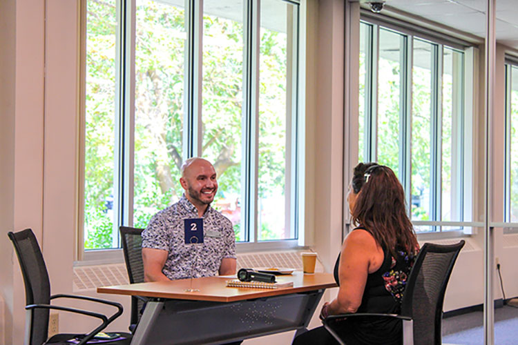Two people seated across from eachother at a desk with large windows in the background.