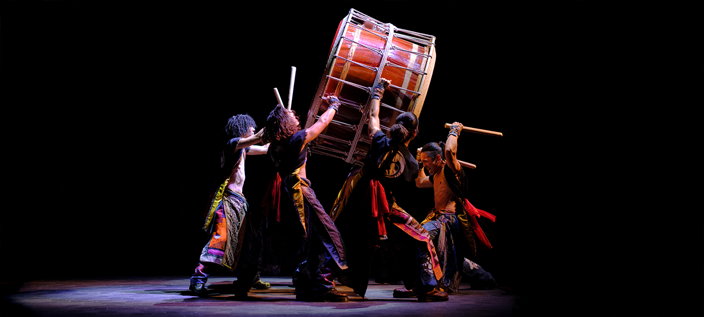 Yamato: The Drummers of Japan performers in a high-energy pose, lifting and striking a large traditional Taiko drum with wooden bachi sticks on a dark stage.