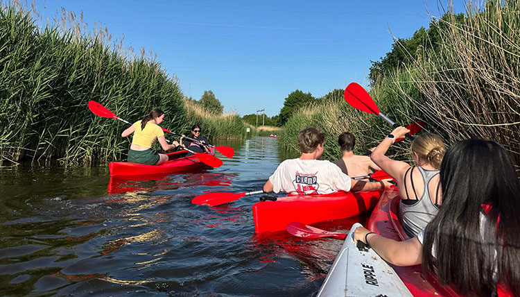 The back sides of three kayaks each with two students haphazardly dipping oars into the water of a grass-lined canal