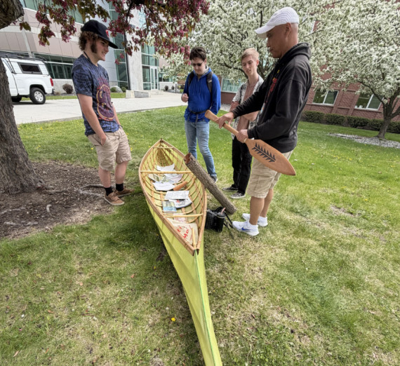 Artist Shawn Brigman, Ph.D., holding a carved wooden paddle while showcasing a handcrafted skin-on-frame canoe to a small group of students on a lawn.