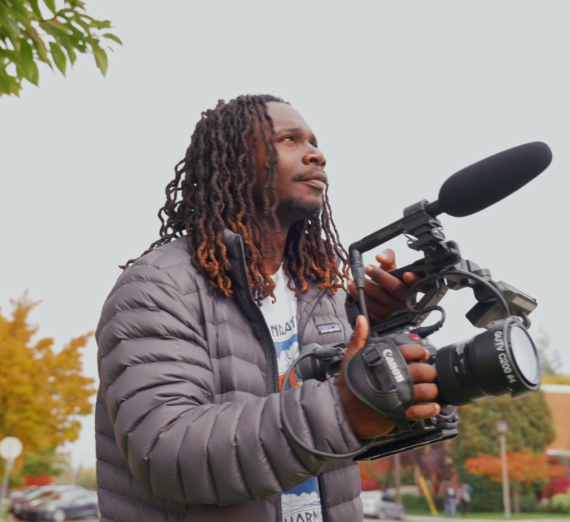 A student standing outside with a video camera filming.