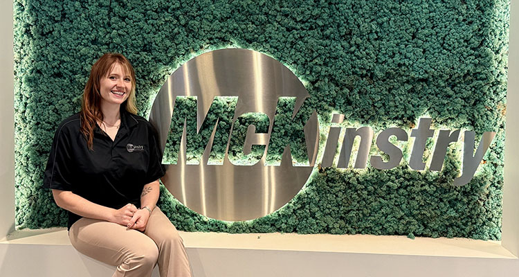 McKinstry Kira Cowell 2026-0723_1352 moss-bd Woman sits in front of a mossy wall with the word McKinstry on it