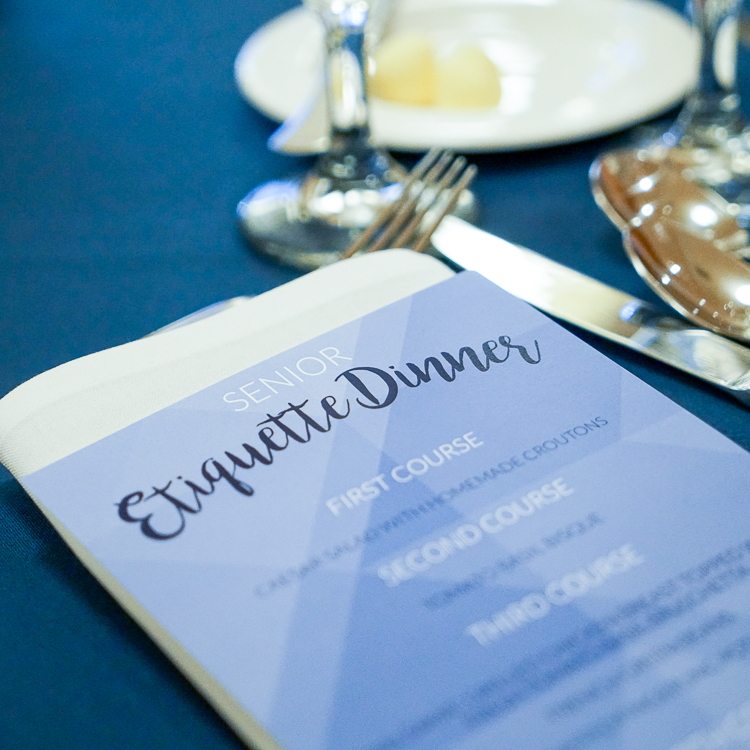 A printed menu rests on a table with a dark blue tablecloth. The menu header reads 'Senior Etiquette Dinner' with the first, second, and third course listed. Silverware is visible just to the left of the menu as well as two stems from wine-style glasses. A printed menu rests on a table with a dark blue tablecloth. The menu header reads 'Senior Etiquette Dinner' with the first, second, and third course listed. Silverware is visible just to the left of the menu as well as two stems from wine-style glasses.
